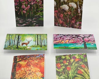 Puede incluir: Seis tarjetas de felicitación con pinturas de paisajes. Las tarjetas presentan varias escenas, incluyendo flores, un ciervo en un bosque y un árbol con un fondo de puesta de sol. Cada tarjeta tiene un interior blanco y se muestra sobre un fondo blanco.