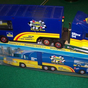 May include: A blue and yellow toy semi-truck with a detachable cab. The truck is labeled "Ultra 94" and "Sunoco Racing Team". The truck is carrying a smaller toy race car. The box the truck is in says "Vehicles in 1 - Detachable Truck Cab - Working Lights - Dual Horns".