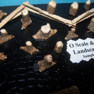 May include: A collection of miniature tree stumps and wooden fence pieces, likely for model train landscaping. The pieces are various sizes and shapes, with a natural wood color. The text on the label reads "O Scale & HO Scale Landscaping Items".