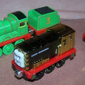 May include: Three toy trains including a green engine with the number 3, a black and gold engine labeled Sodor Ironworks, and a smaller red engine with yellow and black striped details. Each train features a character face on the front.