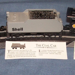 May include: A model train set featuring a yellow Shell tanker car, a gray Shell coal car, and a black steam locomotive. Each car is on a black track with informational cards. The tanker car is yellow with red Shell text.