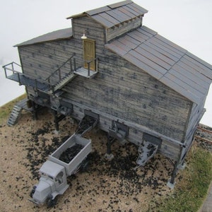 May include: A detailed model of a weathered wooden coal processing facility. The structure features a sloped roof, a loading dock, and a small truck receiving coal. The model is set on a sandy base with scattered coal.