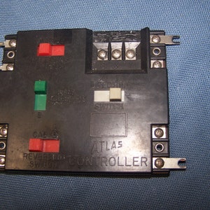 May include: Black Atlas model train controller with red, green, and white switches. The controller has the text "ATLAS CONTROLLER" and "REVERSING SWITCH" printed on it. The controller has metal terminals and is designed for model train use.