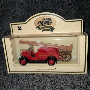 May include: A red commemorative model fire truck with a wooden ladder and a large wooden wheel. The model is in a clear plastic case with a cardboard backing that reads "Made in England". The box has a Chevron logo.