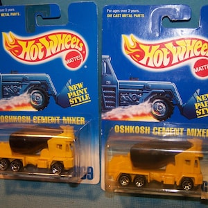 May include: Two Hot Wheels Oshkosh Cement Mixer toy cars on blue packaging. The yellow cement mixers have black drums and silver wheels. The packaging features a blue tractor illustration and the Hot Wheels logo.