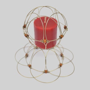 Puede incluir: Una vela roja se encuentra dentro de un portavelas de alambre dorado con una parte superior circular y una base que se asemeja a una flor. El portavelas está decorado con pequeñas cuentas de madera.