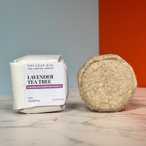 May include: A white bar of shampoo labelled "The Soap Bar" with the text "Lavender Tea Tree" and "For ALL hair types". The bar is wrapped in white paper and is sitting on a white marble surface. A round, grey bar of shampoo is also visible in the image.