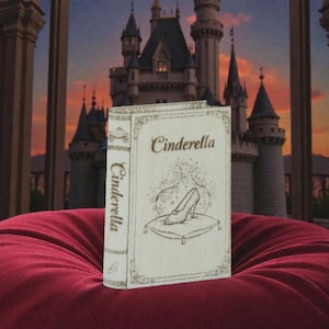May include: A wooden book titled "Cinderella" with a drawing of a glass slipper on a pillow. The book is on a red cushion with a castle backdrop. The book's spine also reads "Cinderella".