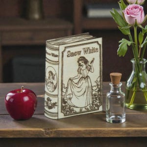 May include: A wooden book-shaped box with the title "Snow White" and an illustration of the character. A red apple, a small glass bottle with a cork stopper, and a vase with pink roses are also present.