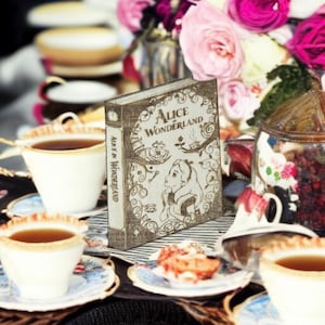 May include: A decorative book titled "Alice in Wonderland" stands upright on a table, next to teacups and a plate of food. The book has a vintage design with illustrations and text. Pink and white flowers are in the background.
