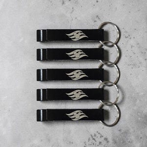May include: Five black keychains with a silver emblem featuring a stylized wave and Mickey Mouse ears. Each keychain has a rectangular shape with a silver key ring attached. The keychains are arranged on a gray surface.