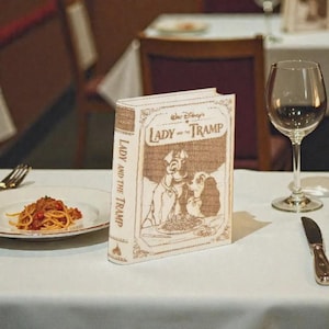 May include: A decorative book with the title "Lady and the Tramp" on the spine and cover, featuring illustrations of the characters. The book is on a white tablecloth next to a plate of spaghetti, a wine glass, and silverware.