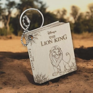 May include: A wooden keychain in the shape of a book, featuring the text "Disney's The Lion King" and an illustration of Simba. The keychain has a silver-colored ring and chain.