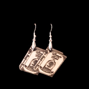 May include: A pair of silver-toned hook earrings featuring rectangular pendants with a light-colored surface. Each pendant displays a black printed illustration of a rose inside a glass dome with the text Beauty and the Beast printed above.