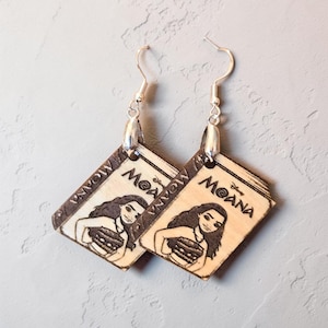 May include: A pair of dangle earrings featuring a light-colored wood finish with a dark, burned-in graphic of the Disney Moana movie logo and the character Moana. The earrings are attached to silver-toned metal fishhook wires for pierced ears.