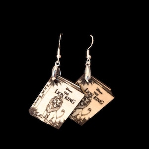 May include: A pair of dangle earrings featuring rectangular wooden charms with a light tan finish. Each charm displays a laser-engraved illustration of a lion and the text Disney's The Lion King. The charms hang from silver-toned metal hooks.