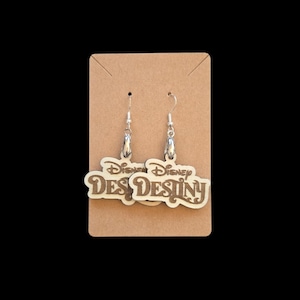 May include: A pair of dangle earrings featuring the word "Disney Destiny" in a stylized font. The earrings are made of light-colored wood with brown lettering and silver-colored ear wires. The earrings are displayed on a brown card.