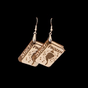 May include: A pair of dangle earrings featuring light-colored wooden book-shaped charms. Each charm is laser-engraved with the word FROZEN at the top, a silhouette of a person with long hair, and small snowflake-like patterns. Silver-toned hooks.