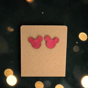 May include: A pair of red wooden stud earrings in the shape of a mouse head, displayed on a brown card. The earrings have a smooth finish and are set against a dark green background with bokeh lights.