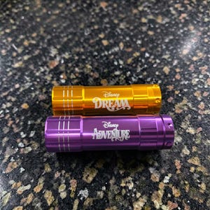 May include: Two small flashlights, one gold and one purple, with the Disney logo and the words "Dream" and "Adventure" printed on them. The flashlights are cylindrical and have vertical lines etched on the sides.