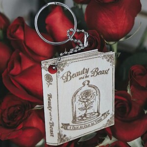May include: Wooden keychain featuring the Beauty and the Beast book cover design. The cover shows a rose in a glass dome, with the text "Beauty and the Beast" and "A Tale as Old as Time". The keychain is attached to a silver keyring.