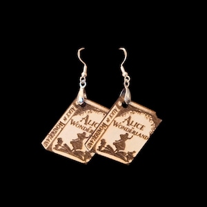 May include: A pair of light-colored wooden earrings shaped like miniature books. Each earring features a laser-engraved illustration of a character and the text Alice in Wonderland on the front, suspended from silver-toned metal fishhook wires.