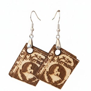 May include: A pair of dangling earrings featuring square, light-brown wooden charms laser-engraved with a silhouette of a mermaid on a rock and the text The Little Mermaid. The charms hang from silver-toned metal fishhook ear wires.