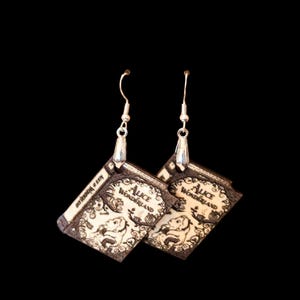 May include: A pair of dangle earrings featuring miniature book-shaped charms. Each charm is cream-colored with dark brown detailing, depicting a character and the text Alice in Wonderland on the cover. The charms hang from silver-toned hooks.