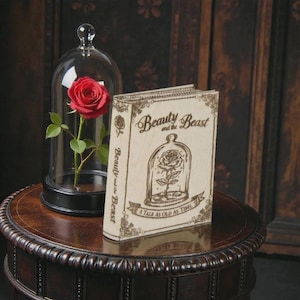 May include: A red rose in a glass cloche and a decorative book. The book cover reads "Beauty and the Beast" with an illustration of a rose in a glass dome. The book is on a dark brown table with a decorative edge.