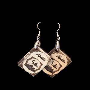 May include: A pair of dangling earrings featuring miniature book-shaped charms made of light-colored wood with dark brown edges. The front of each charm is engraved with the word Aladdin, a crescent moon, and a silhouette of a flying carpet.