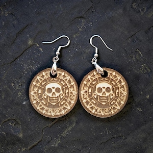 May include: A pair of wooden earrings featuring a skull design within a circular pattern. The earrings have silver-colored ear wires. The wood has a light brown color, and the design is etched into the surface.