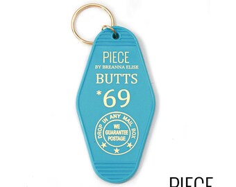 Può includere: Un portachiavi blu a forma di chiave di motel con un anello di chiave dorato. Il portachiavi ha il testo "PIECE BY BREANNA ELISE BUTTS *69" e un cerchio con il testo "DROP IN ANY MAIL WE GUARANTEE POSTAGE BOX".