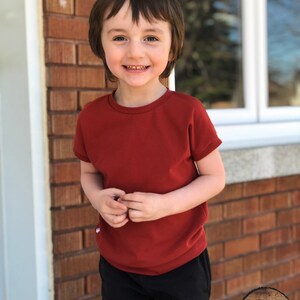 May include: A young child wearing a red short-sleeved t-shirt and black pants. The child is smiling and looking at the camera. The background is a brick wall.