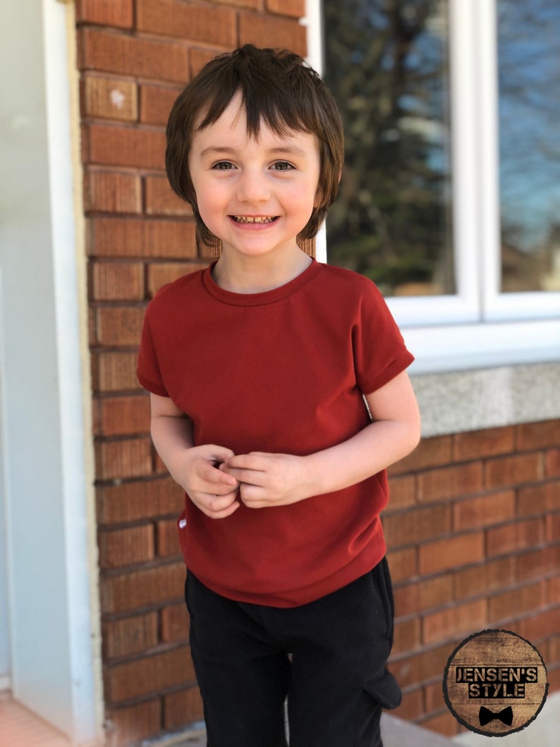 May include: A young child wearing a red short-sleeved t-shirt and black pants. The child is smiling and looking at the camera. The background is a brick wall.