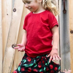 May include: A young girl wearing a red short-sleeved shirt and a black skirt with a cherry print. The skirt has a white lining and is gathered at the waist.