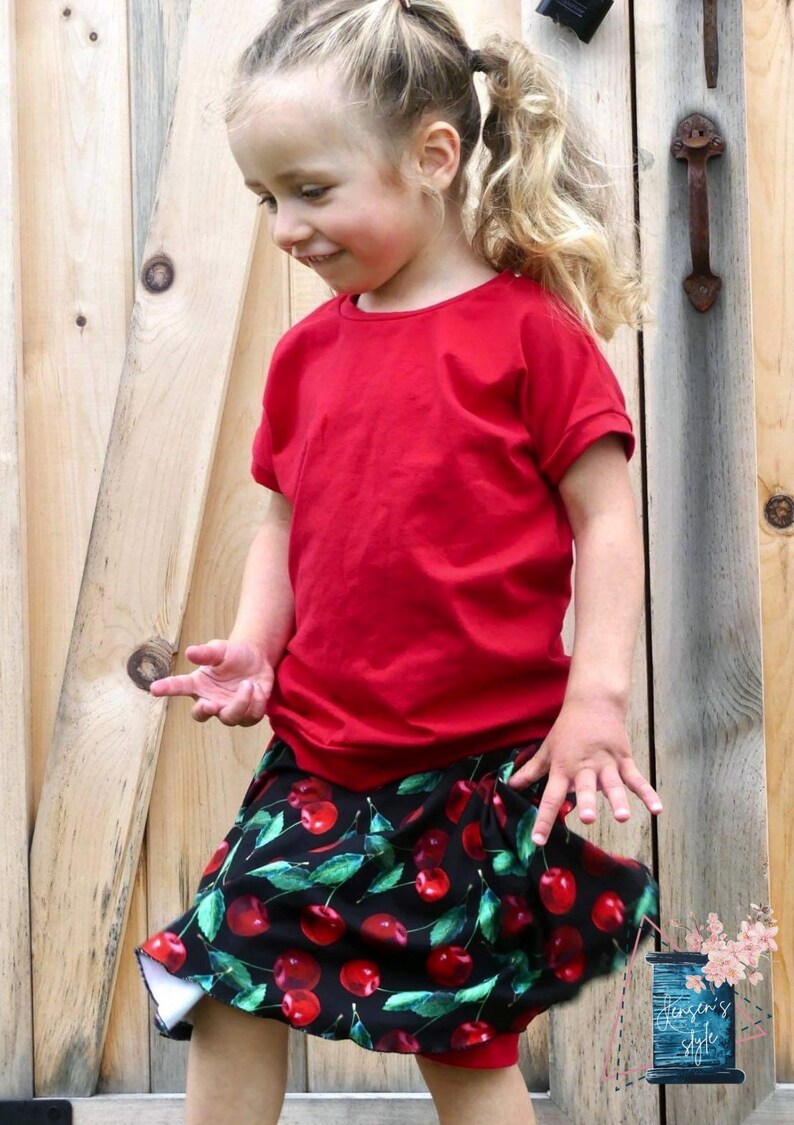 May include: A young girl wearing a red short-sleeved shirt and a black skirt with a cherry print. The skirt has a white lining and is gathered at the waist.
