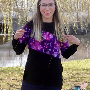 Op de afbeelding: Een vrouw draagt een zwart en paars langarm shirt met een galaxy print. Het shirt heeft een diagonaal ontwerp met een zwart gedeelte aan de onderkant. De vrouw draagt een blauwe jeans en heeft lang bruin haar.