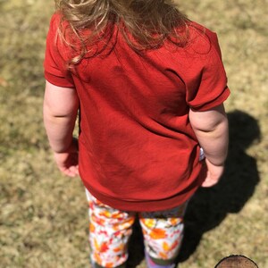 May include: A young child wearing a red short-sleeved shirt and colorful patterned pants. The child is standing on a grassy surface.