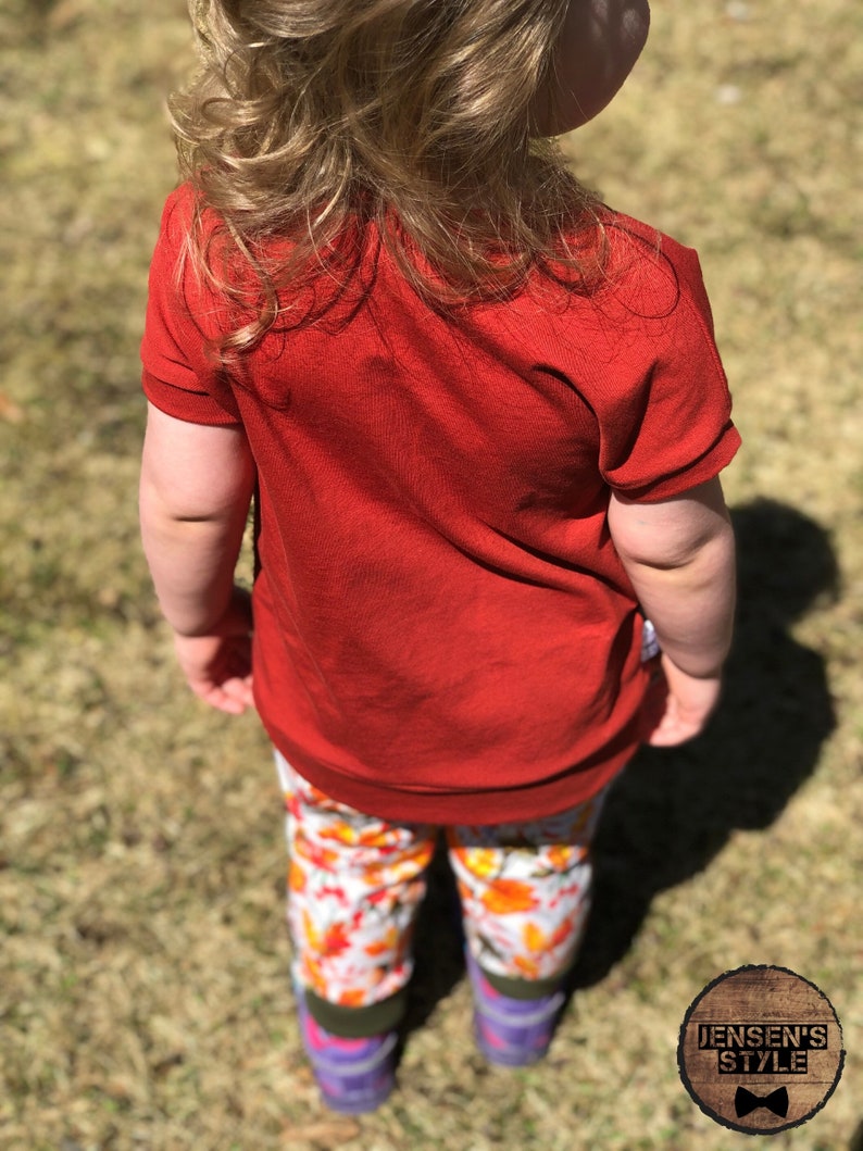 May include: A young child wearing a red short-sleeved shirt and colorful patterned pants. The child is standing on a grassy surface.