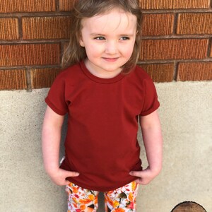 May include: A young child wearing a red short-sleeved shirt and white pants with a fall leaf print. The child is standing in front of a brick wall.