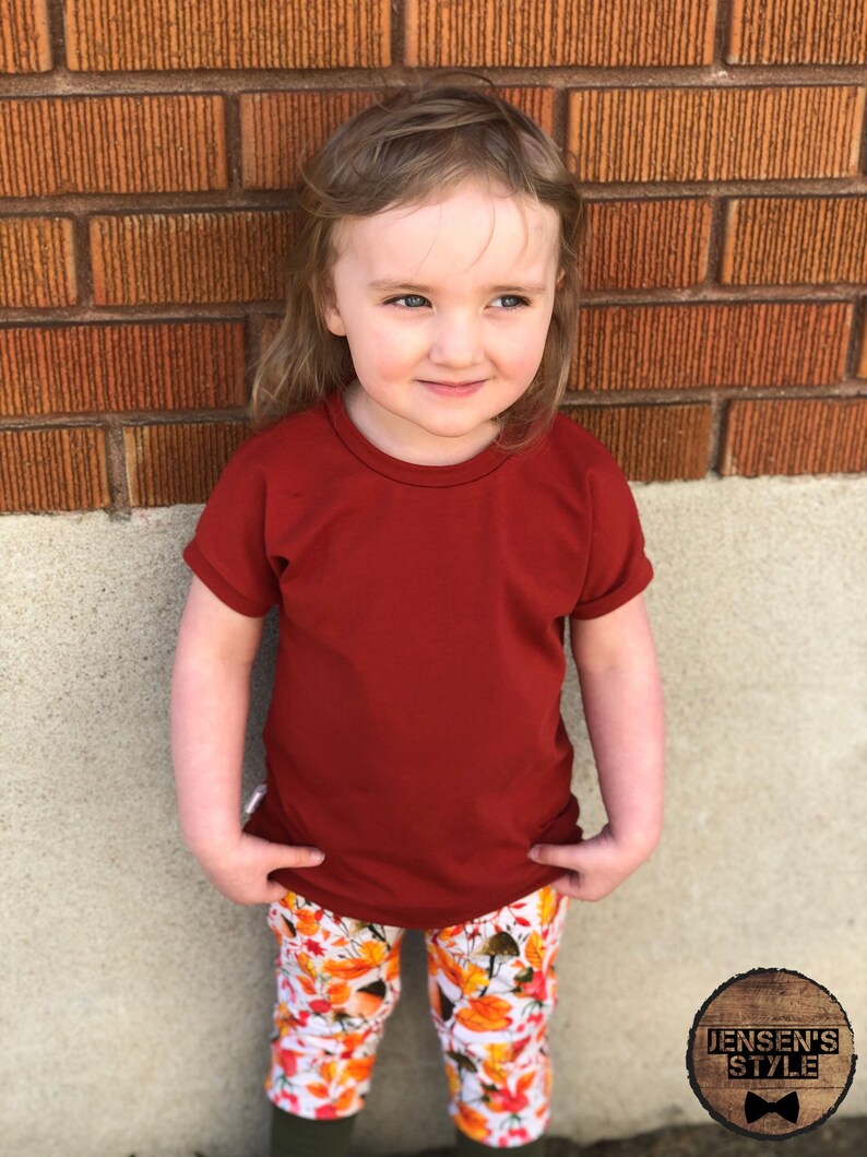 May include: A young child wearing a red short-sleeved shirt and white pants with a fall leaf print. The child is standing in front of a brick wall.