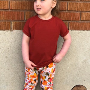 May include: A young child wearing a red short-sleeved shirt and white leggings with a fall leaf print. The leggings have a brown, orange, and yellow leaf pattern. The child is standing in front of a brick wall.