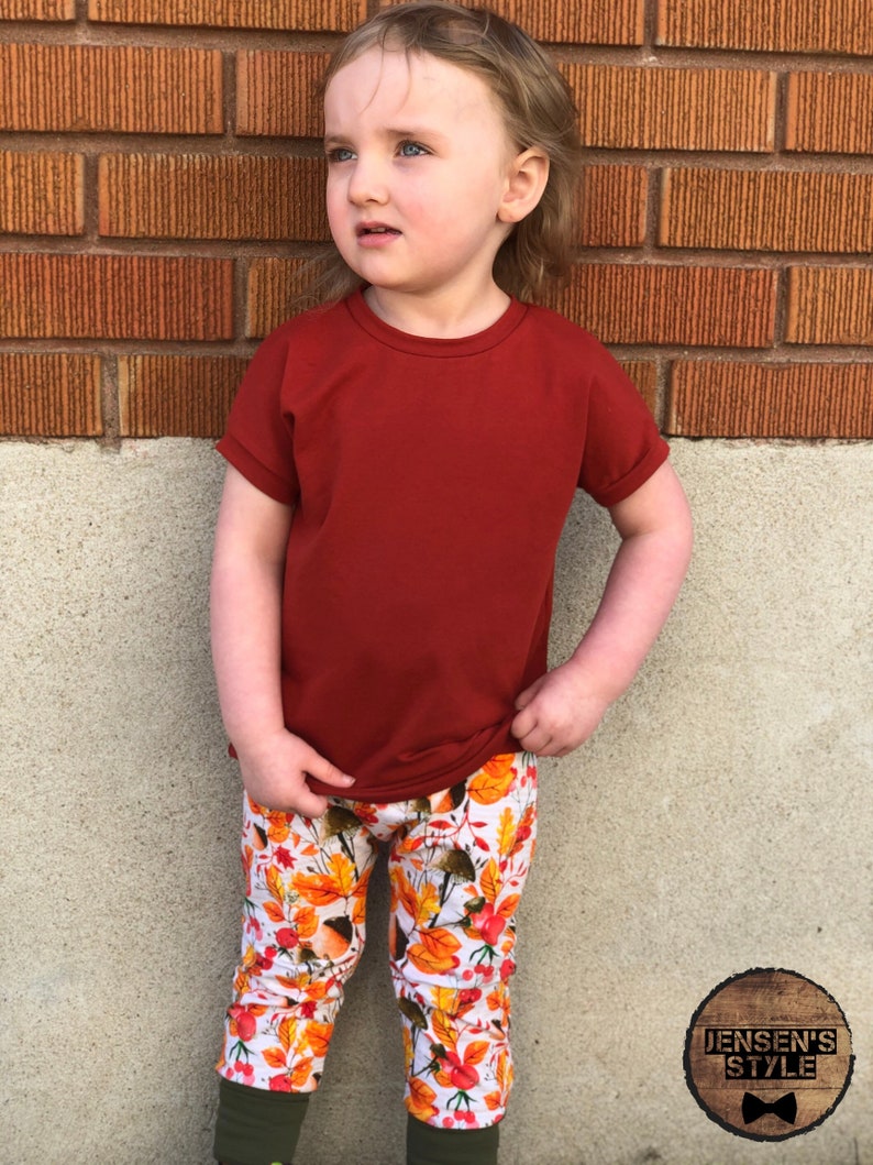 May include: A young child wearing a red short-sleeved shirt and white leggings with a fall leaf print. The leggings have a brown, orange, and yellow leaf pattern. The child is standing in front of a brick wall.
