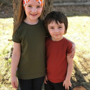 May include: Two children, a girl wearing a green short-sleeved shirt and a headband with orange and white flowers, and a boy wearing a red short-sleeved shirt, are standing next to each other. They are both smiling and looking at the camera. The girl has her arm around the boy's shoulder. The background is a blurry green lawn and a tree trunk.