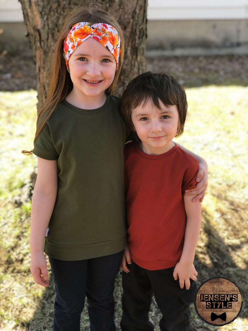 May include: Two children, a girl wearing a green short-sleeved shirt and a headband with orange and white flowers, and a boy wearing a red short-sleeved shirt, are standing next to each other. They are both smiling and looking at the camera. The girl has her arm around the boy's shoulder. The background is a blurry green lawn and a tree trunk.