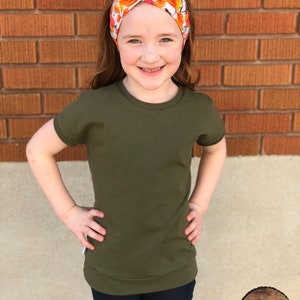 May include: A young person wearing a green short-sleeved shirt with a fall leaf headband. The shirt has a relaxed fit and the headband is made of fabric with a white background and orange and yellow leaves.