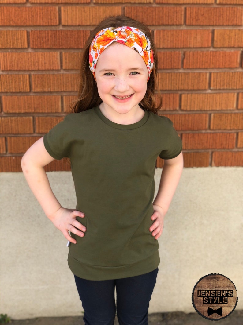May include: A young person wearing a green short-sleeved shirt with a fall leaf headband. The shirt has a relaxed fit and the headband is made of fabric with a white background and orange and yellow leaves.