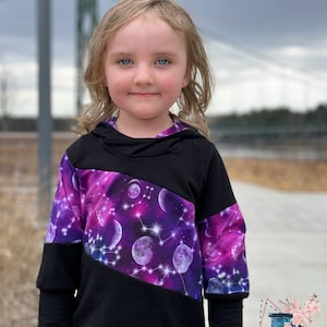 May include: A black and purple hooded sweatshirt with a celestial galaxy design. The sweatshirt features a black hood and sleeves, with a diagonal panel of purple fabric showcasing planets, stars, and constellations. The design is for children.