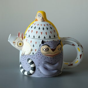 Può includere: Una teiera fantasiosa con un lemure viola, un coniglio bianco e una donna con i capelli gialli sul coperchio. La teiera è decorata con cuori rossi e picche nere.