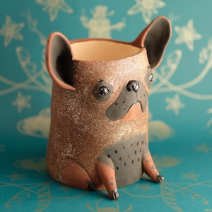 Puede incluir: Un macetero de cerámica marrón y negro en forma de bulldog francés sentado, con detalles de purpurina. El macetero tiene la forma de un perro sentado con la nariz y los ojos negros.